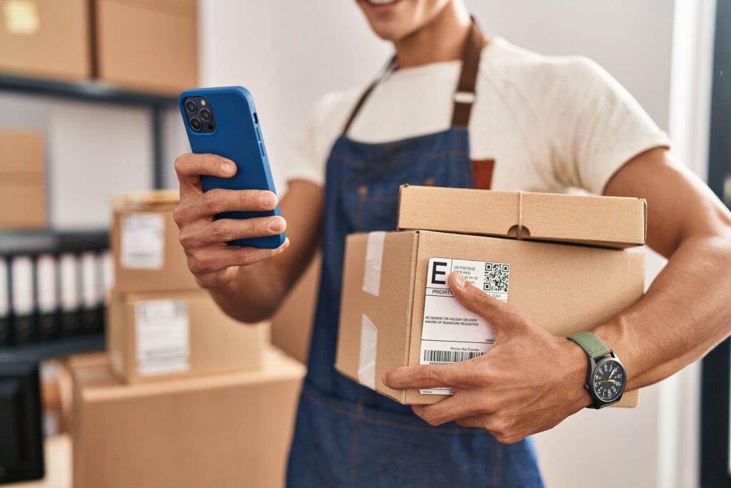 Young hispanic man ecommerce business worker holding packages and using smartphone at office